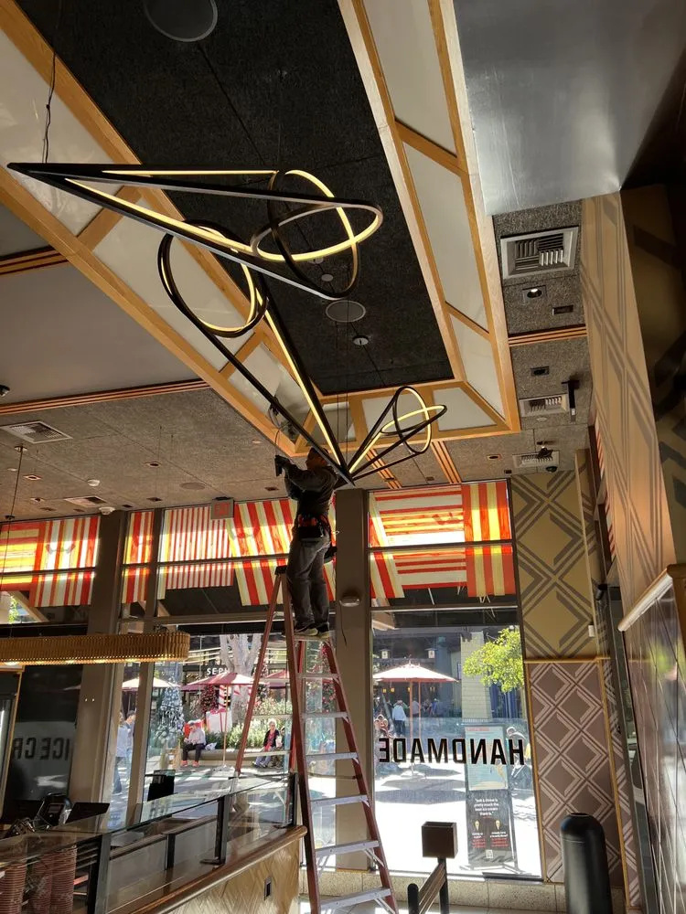 Electrician on a ladder installing a sculptural LED pendant in a restaurant storefront.